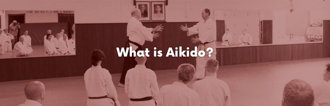 White text that reads “What is Aikido?“ overlaid on a photo of people participating in an aikido class. 